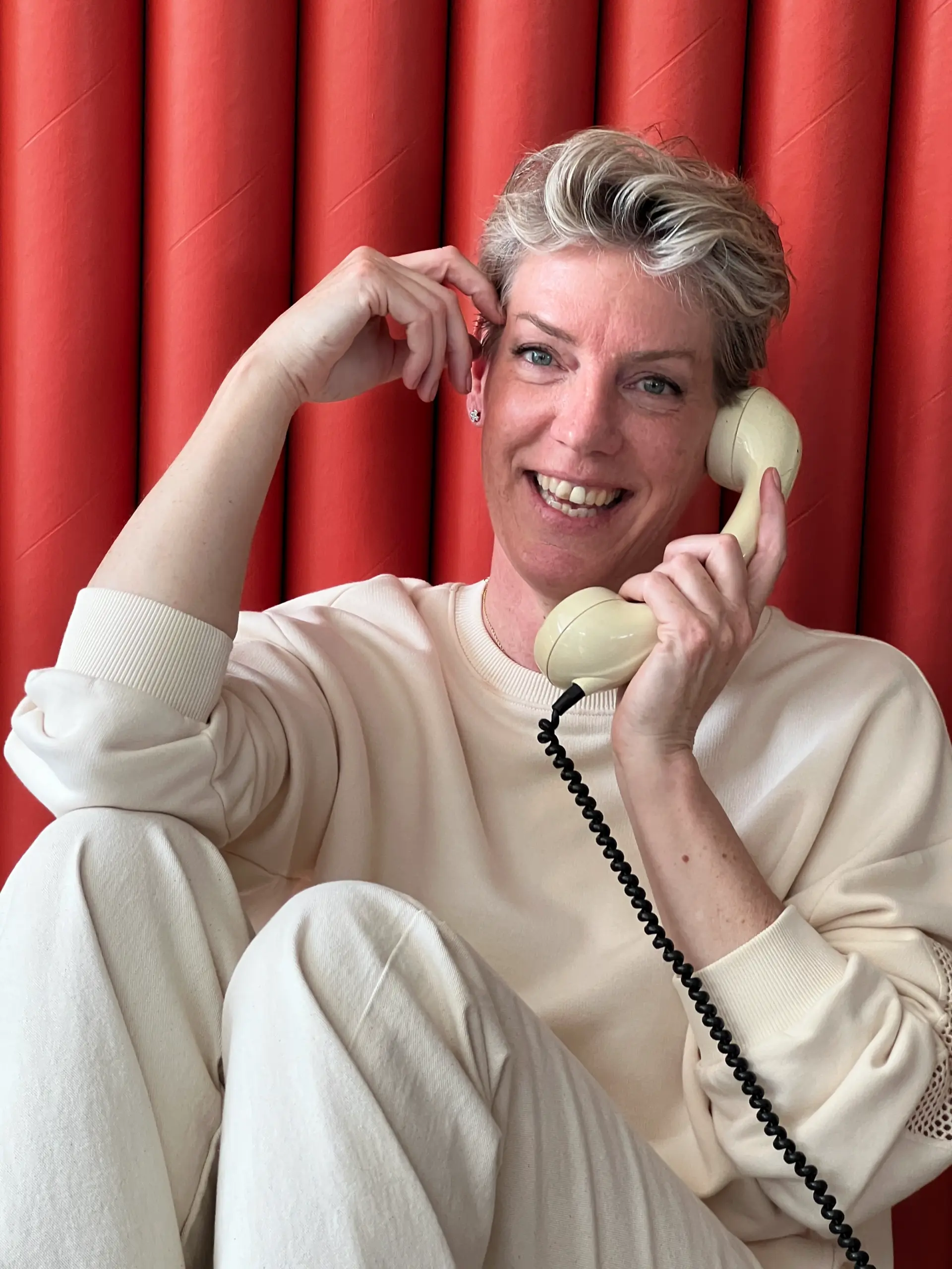 Kirsten Hermsen lacht en belt met een crèmekleurige telefoon voor een rood gordijn