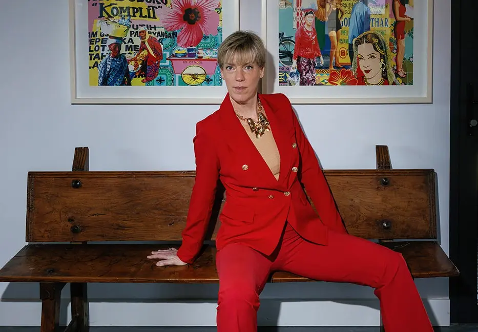 Kirsten Hermsen in een rood pak op een houten bank onder kleurrijke kunst